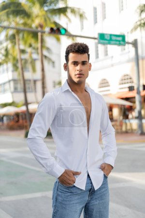 gut aussehender kubanischer Mann in weißem Hemd und Jeans mit Händen in Taschen auf verschwommener Straße in Miami