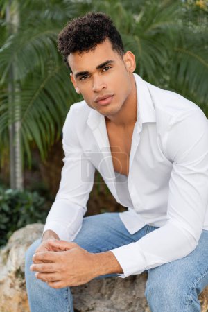 schöner kubanischer Mann in weißem Hemd und Jeans, der in die Kamera schaut, während er auf einem Stein in der Nähe von Palmen sitzt