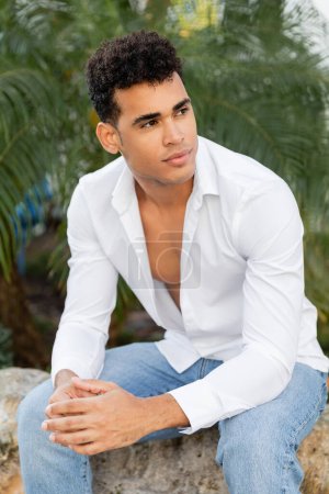 Porträt eines gutaussehenden und kubanischen Mannes in weißem Hemd und Jeans, der auf Stein sitzend wegschaut