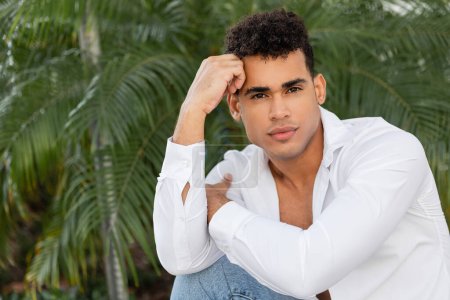 Gut aussehender junger kubanischer Mann mit lockigem Haar in weißem Hemd und Jeans posiert in der Nähe von Palmen 