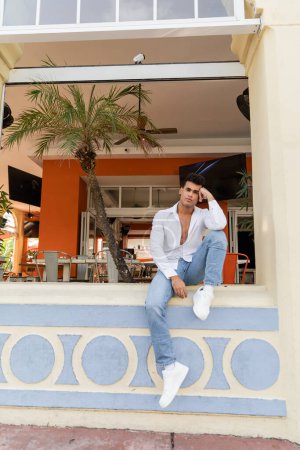 young cuban man in white shirt and jeans sitting on parapet of outdoor cafe with palm trees in Miami