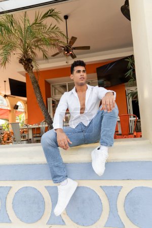 Téléchargez les photos : Homme cubain en chemise blanche et jeans assis sur le parapet d'un café en plein air avec des palmiers à Miami - en image libre de droit