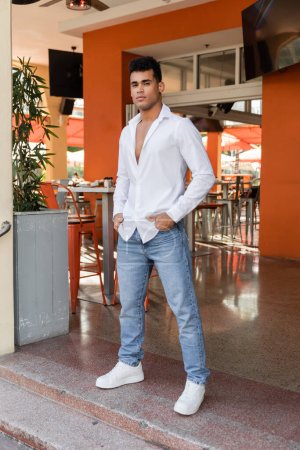 Longitud completa del hombre cubano en camisa blanca y jeans posando en cafetería al aire libre en la calle urbana de Miami