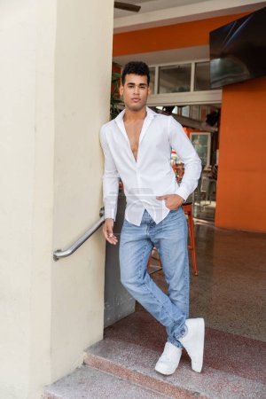 Longitud completa del hombre cubano en camisa blanca y jeans posando en cafetería al aire libre en la calle en Miami