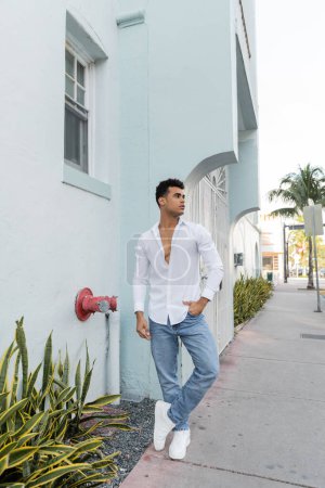 Full length of curly cuban man in stylish shirt and blue jeans posing on street in Miami