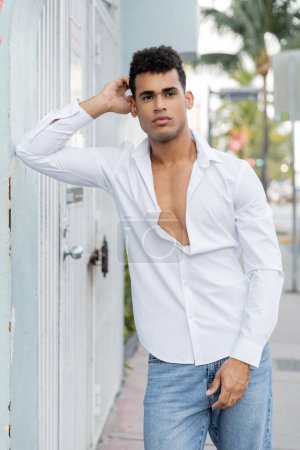 curly young cuban man in stylish shirt and blue jeans posing on street in Miami