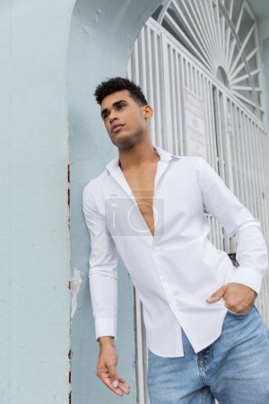 schöner und lockiger kubanischer Mann in stylischem Hemd und blauer Jeans posiert auf der Straße in Miami