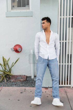 Longitud completa de un joven cubano relajado mirando hacia otro lado mientras está de pie cerca del edificio en la calle en Miami