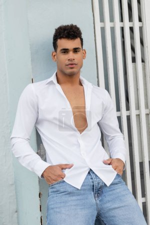 Modischer und gut aussehender kubanischer Mann in Hemd und Jeans posiert in der Nähe eines Gebäudes in Miami