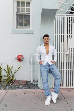 Ein trendiger kubanischer Mann in Hemd und Jeans steht in voller Länge in der Nähe eines Gebäudes mit Tor in Miami