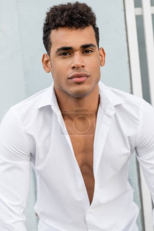 Porträt eines lockigen und stylischen kubanischen Mannes im weißen Hemd, der im Urlaub in Miami in die Kamera schaut