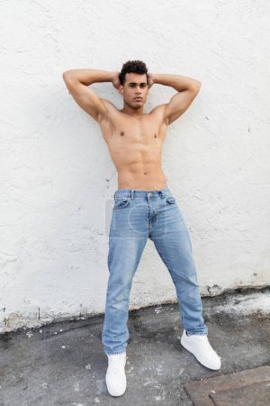 Full length of athletic cuban man in blue jeans posing near building on  street in Miami