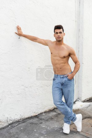Full length of strong and shirtless young cuban man in blue jeans posing and touching wall in Miami 