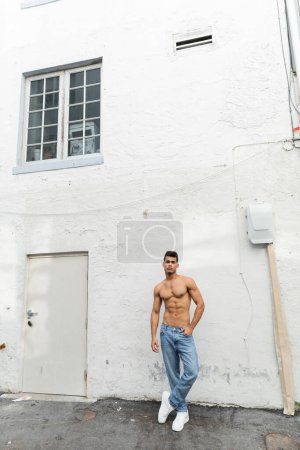 Full length of muscular and shirtless cuban man in blue jeans posing near building in Miami