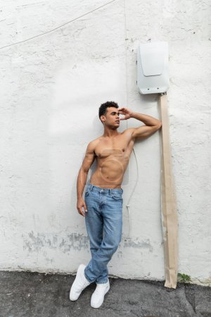Shirtless cuban man in jeans standing near wire on white wall in Miami during summer, muscular 