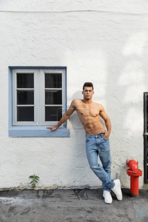 Full length of sexy and shirtless young cuban man in eyeglasses and blue jeans in Miami