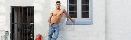 Sexy y sin camisa joven hombre cubano en pantalones vaqueros azules y gafas de pie cerca del edificio, pancarta