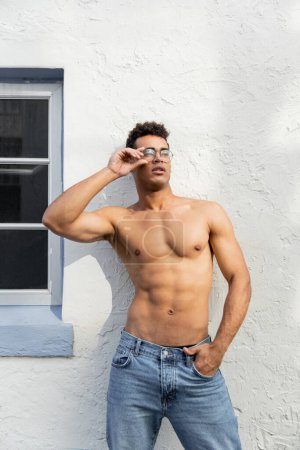 shirtless cuban man in round-shaped eyeglasses holding hand in pocket of jeans, standing in Miami