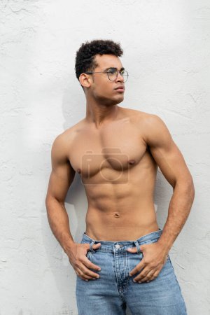 athletic young cuban man in jeans and round-shaped eyeglasses looking away, standing near white wall 
