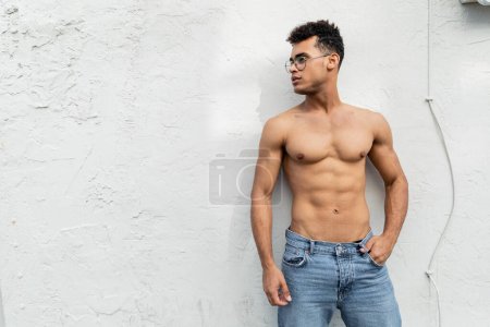 homme cubain avec corps athlétique posant en jeans bleus et des lunettes de vue à la mode en forme ronde près du bâtiment