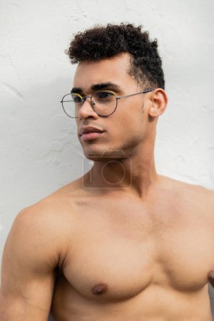 Portrait of shirtless and muscular young cuban man in stylish round-shaped eyeglasses looking away