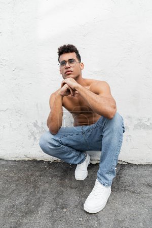 Shirtless and young cuban man with athletic body wearing round-shaped eyeglasses and blue jeans