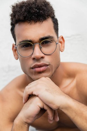 Portrait of curly and shirtless cuban man in stylish round-shaped eyeglasses holding hands near chin