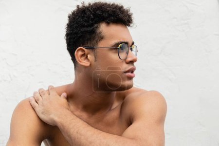 Young shirtless cuban man in trendy round-shaped eyeglasses touching shoulder and looking away