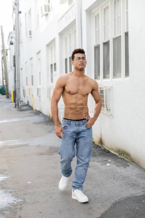 sexy cuban man in round-shaped eyeglasses and jeans looking away while walking in Miami