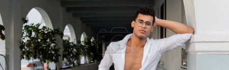 Sexy young cuban man in stylish eyeglasses and white shirt looking at camera in Miami, banner