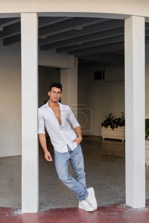 Foto de Longitud completa del hombre cubano con estilo en gafas redondas, camisa blanca y jeans de pie en Miami - Imagen libre de derechos