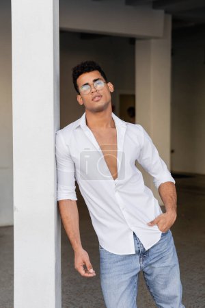 Trendy young cuban man in round-shaped eyeglasses and white shirt holding hand in pocket 