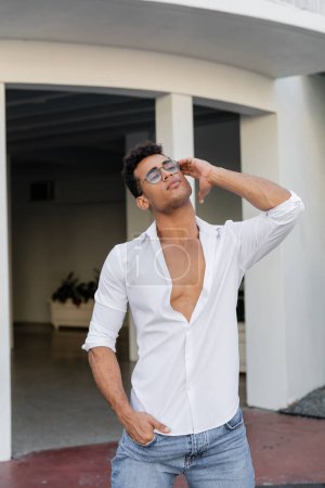 Relaxed and good looking cuban man in white shirt and round-shaped eyeglasses touching curly hair 