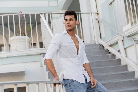 Téléchargez les photos : Good looking cuban man in white shirt and jeans looking away while standing near railing in Miami - en image libre de droit