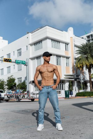 sexy und junger kubanischer Mann mit Baseballmütze hält die Hände in den Taschen auf einer städtischen Straße in Miami