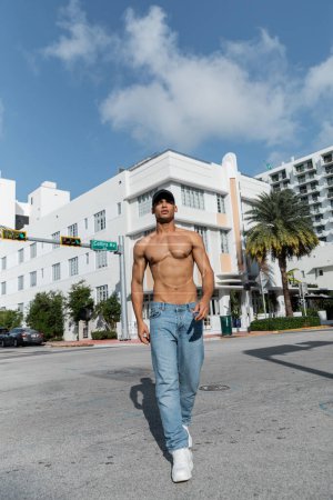shirtless young cuban man in jeans and baseball cap walking on urban street in Miam, during summer 