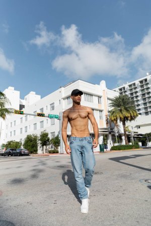 shirtless young cuban man in jeans and baseball cap walking on urban street in Miam, summer 