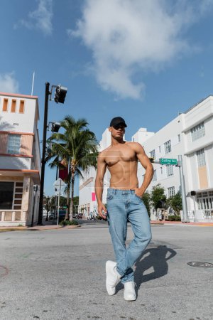 jeune homme cubain avec corps athlétique en casquette de baseball et jeans bleus dans la rue à Miami, plage sud
