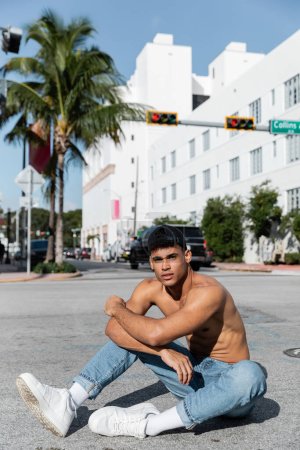 sexy cuban man in baseball cap and blue jeans sitting on road in Miami 