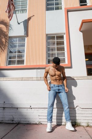 Téléchargez les photos : Homme cubain torse nu avec corps athlétique en casquette de baseball et jeans debout dans la rue à Miami - en image libre de droit