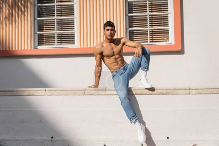 Téléchargez les photos : Homme cubain avec corps musclé posant dans casquette de baseball et jeans dans la rue à Miami en été - en image libre de droit