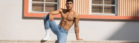 Foto de Hombre cubano con cuerpo musculoso posando en gorra de béisbol y jeans en la calle en Miami en verano, pancarta - Imagen libre de derechos