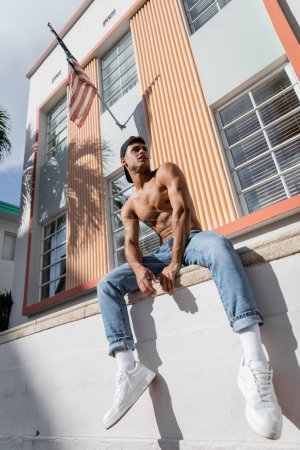 Téléchargez les photos : Homme cubain en casquette de baseball, jeans et baskets assis sur le parapet de la construction avec drapeau américain - en image libre de droit