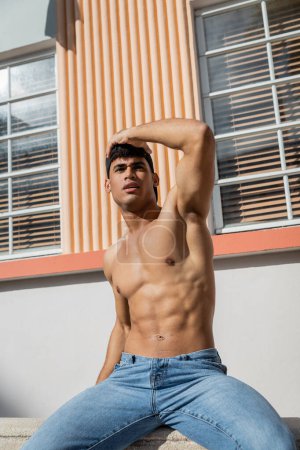 Téléchargez les photos : Athlète jeune homme cubain en casquette de baseball et jeans bleus regardant loin tout en étant assis sur parapet - en image libre de droit