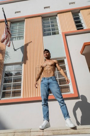 hombre cubano musculoso en gorra de béisbol y jeans de pie en parapeto cerca del edificio con bandera americana 