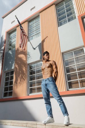 Shirtless cuban man in baseball cap and jeans standing on parapet near building with american flag 