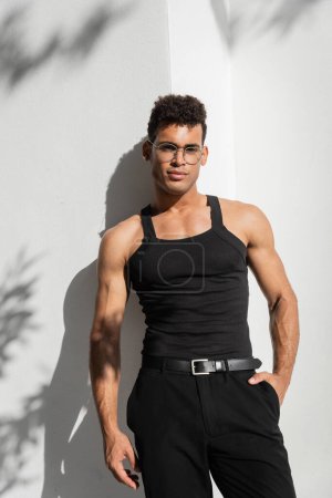 Téléchargez les photos : Portrait d'homme cubain musclé à la mode en lunettes et t-shirt noir sans manches posant près du mur - en image libre de droit
