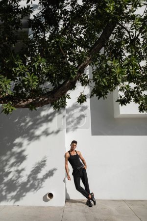 Elegante joven cubano en traje negro y anteojos posando cerca del edificio con sombra
