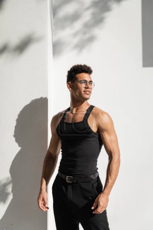 Téléchargez les photos : Élégant et joyeux jeune homme cubain dans des lunettes près du bâtiment avec ombre sur la rue à Miami - en image libre de droit