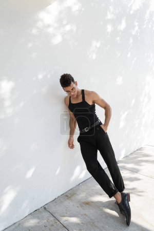 Téléchargez les photos : Homme cubain en lunettes et tenue noire tenant la main dans la poche du pantalon et debout près du mur - en image libre de droit